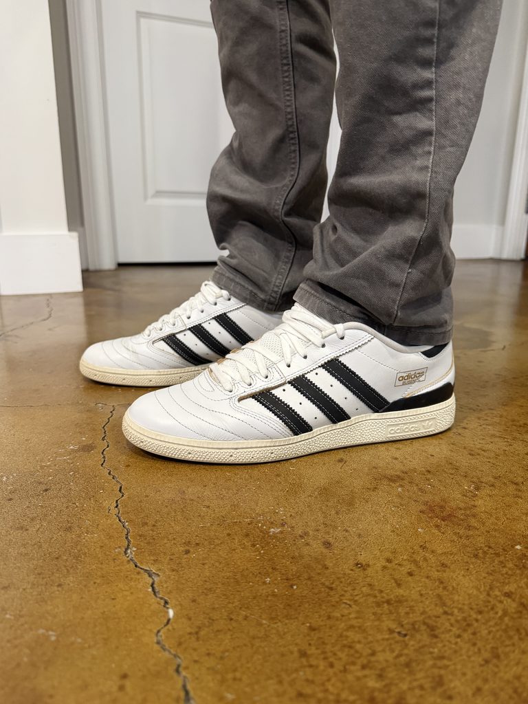 Close-up on-feet view of white leather adidas Busenitz sneakers with three black-stripes and an off white sole, styled with relaxed-fit grey pants.